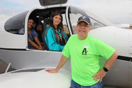 People seated in a small airplane cockpit while a man in an Airport Day shirt stands beside the plane.