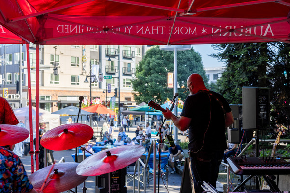 A guitarist performs on an outdoor stage as people gather at tables in downtown Auburn.