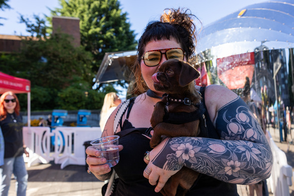 A tattooed woman holds a small brown puppy at an outdoor event.
