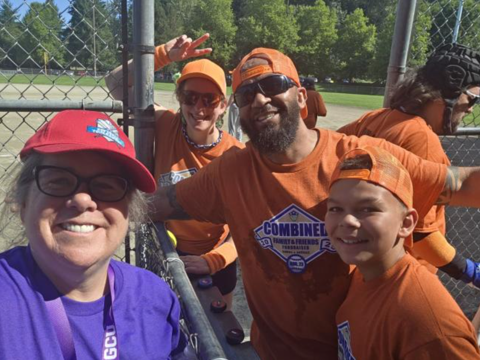 a woman takes a selfie with four people in orange shirts at a softball field.