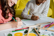 Two people draw and paint together at a table covered with art supplies.