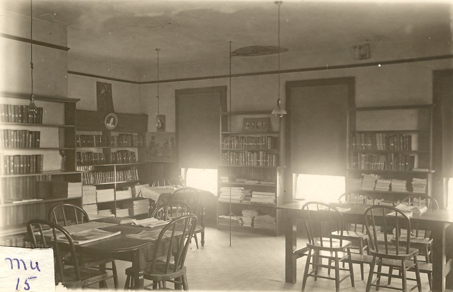 Historic black-and-white photo of a library reading room with bookshelves, tables, chairs and stacks of books.