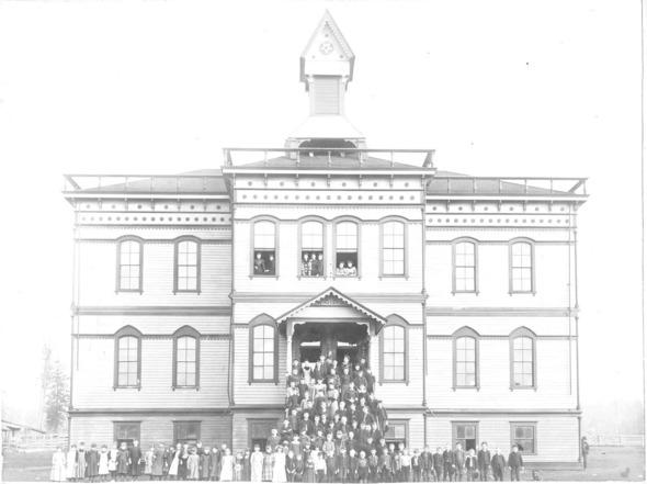 Historic black-and-white photo of students gathered outside a two-story wooden schoolhouse with a central bell tower.