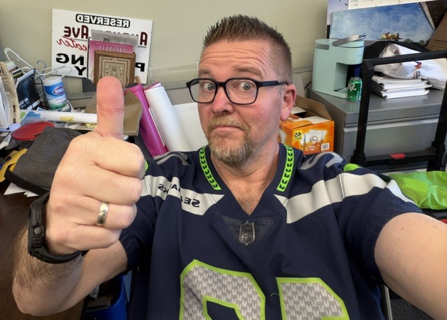 Man wearing a Seahawks jersey gives a thumbs up while seated in an office.