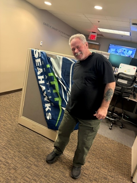 Man smiles beside a Seahawks flag displayed in an office cubicle.