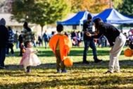 A child in a pumpkin costume and a toddler in a pink dress pose for a photo at an outdoor fall festival.