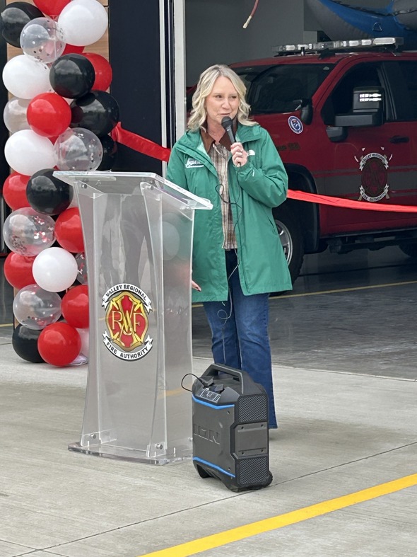 A woman in a green jacket speaks at a VRFA podium beside a fire engine and red, white and black balloons.