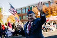 Smiling veteran in uniform cap waves while carrying two American flags during Auburn’s Veterans Parade.
