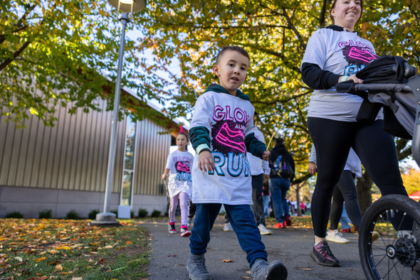 Young child in a Glow Run shirt walks along a tree-lined path beside an adult pushing a stroller.