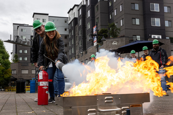 Two people in green hard hats practice using a fire extinguisher on a live training fire as others watch.