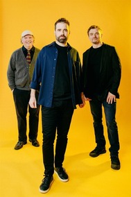 Three members of the band Duende Libre pose against a bright yellow background.