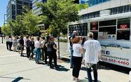 Image shows people lined up in front of a food truck. The menu and window for the truck is visible. 