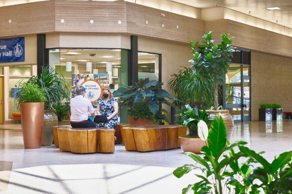 Indoor area at Crossroads Mall with wooden benches, large potted plants, and two people seated at a table near a “Welcome to Mini City Hall” sign.