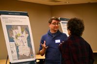 A person speaks with another attendee in front of large display boards showing maps and project information at a public event.