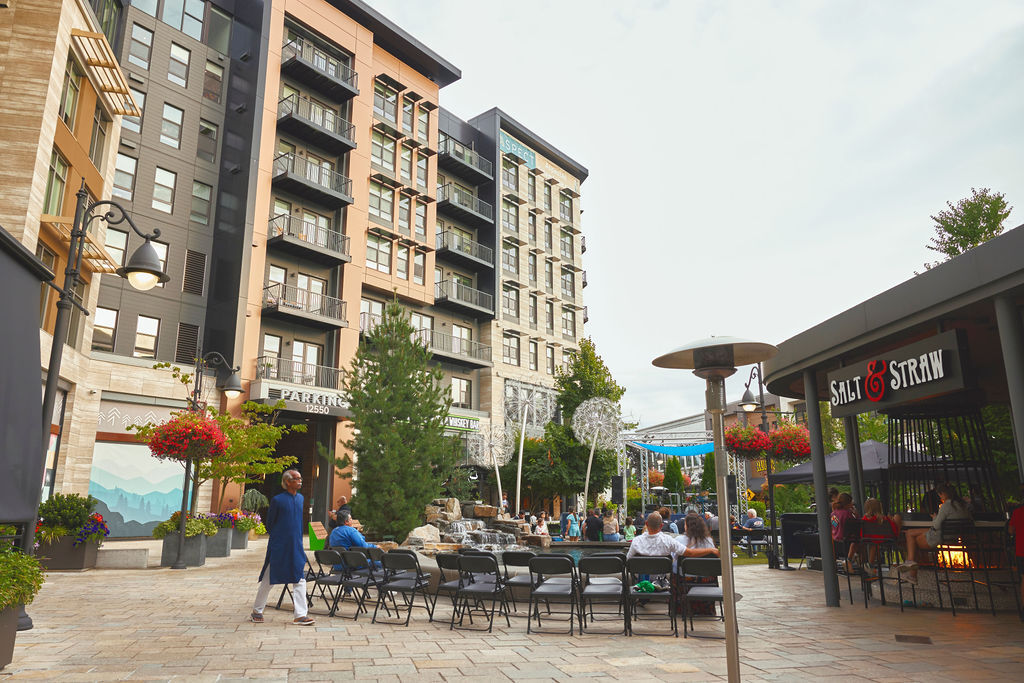 Outdoor urban plaza with mid‑rise buildings, seating, landscaping, and people gathered near shops and a small fountain.