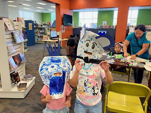 Two children wear handmade masks while families make masks in the background