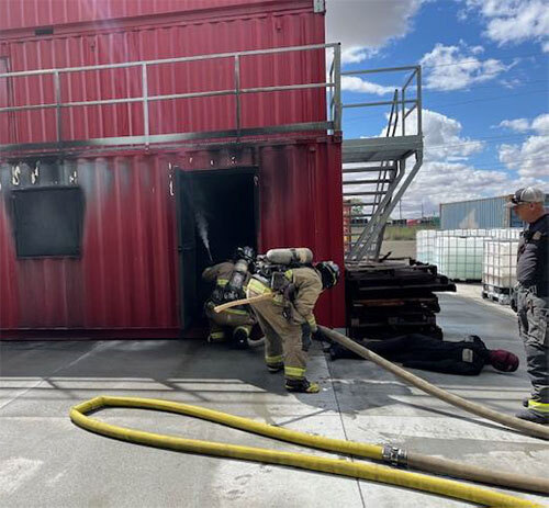 Two firefighters hold a hose and spray water through a doorway into a training tower