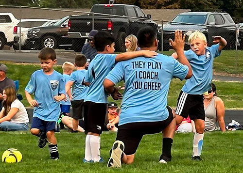 An adult soccer coach gives a high-five to a young player with other players and family members around them