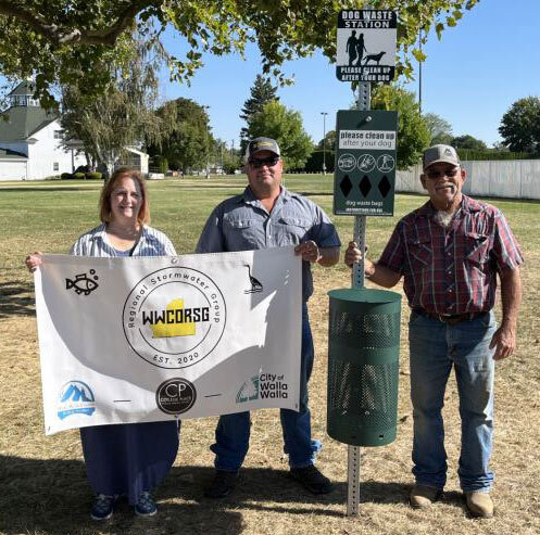 Three people stand with a pet waste station and a banner at the Walla Walla County Fairgrounds