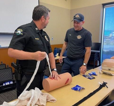 Using a fake body part, a fire department employee shows a police department employee how to bandage a wound