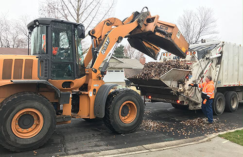 Front-loader dumps leaves into the back of a garbage truck while a crew member holds a control arm