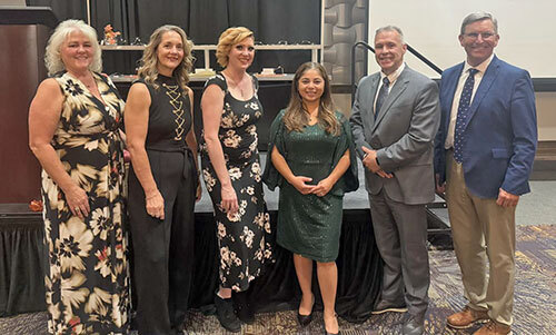 Six standing people pose for a group photo in a ballroom