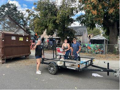 Three people pause for a photo while moving items from a trailer to a dumpster on a residential street