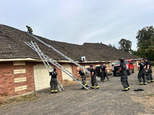 Firefighters wearing turnout pants and T-shirts practice putting ladders on a house roof and climbing up them
