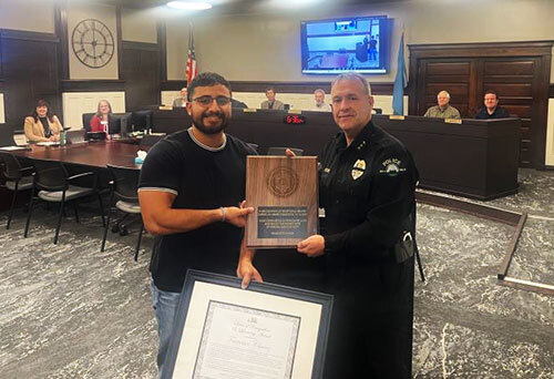 A man in police uniform and another man pose for a photo in City Council Chambers while holding a framed certificate and a plaque