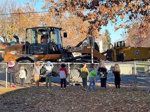 Children stand by a chain-link fence and watch a front-loader picking up leaves in the street