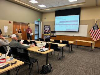 People sit at tables in front of a video screen while two people present information