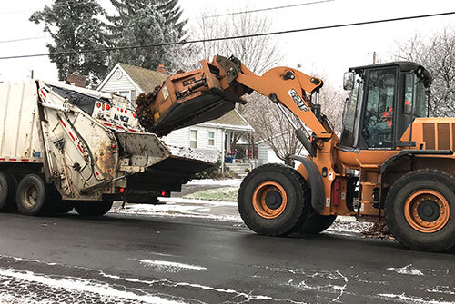 Worker in a front-loader uses a scoop to drop leaves into the back of a garbage truck