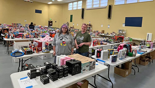 Two Toys For Tots volunteers stand in a gym with tables covered with toys and games