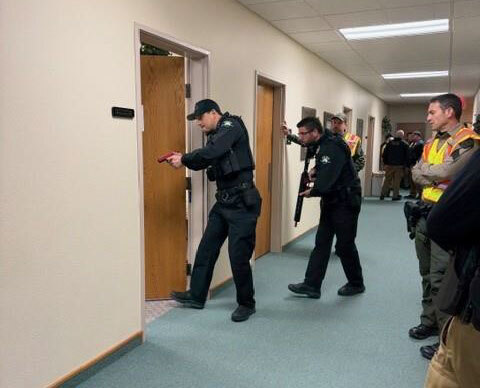 Police officers holding firearms train in the hallway of a building while other personnel observe