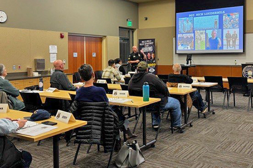 Attendees of the Citizens Academy sit at tables while a police officer gives a presentation.