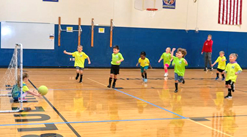 Nine children play soccer in a gymnasium while an adult coach looks on