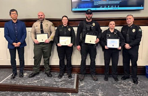 A mayor and five law enforcement personnel — four holding awards — stand in the Council Chambers