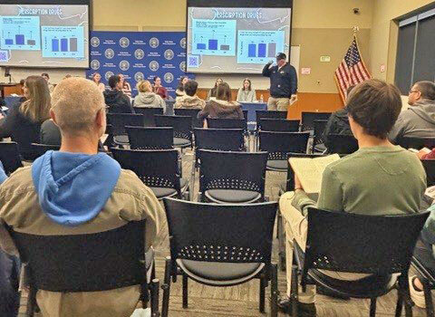 People sit in a room and watch a presentation by a law enforcement officer standing by a panel of youths
