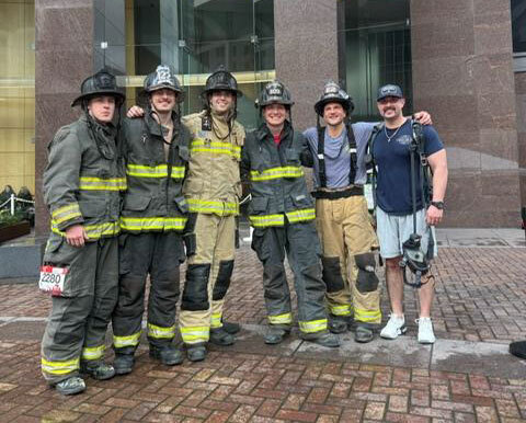Six firefighters in gear stand in front of a building