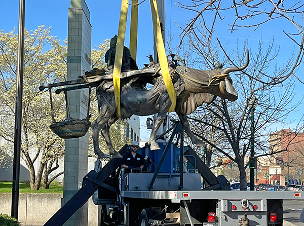 The statue of Matilde the cow is suspended by yellow straps as a crane moves it onto a trailer