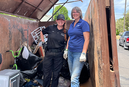 Two women wearing gloves stand in the doorway of a large dumpster during a neighborhood cleanup event