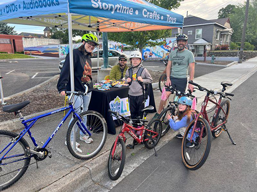Two parents and two children stand with bicycles next to a canopy outside the library with a table that has prizes on it