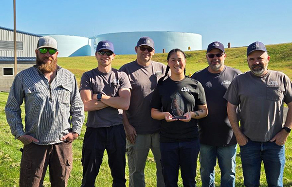 Six Water Division employees, one holding a trophy, stand in front of two blue water storage facilities