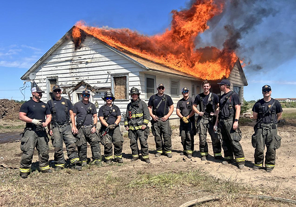 Ten firefighters in turnout gear stand outdoors in front of a small home with flames coming off its roof