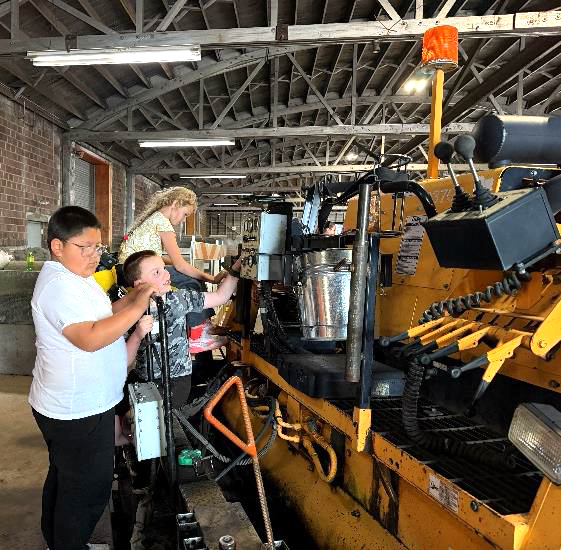 Three elementary students explore a piece of paving equipment in a Street Division shop
