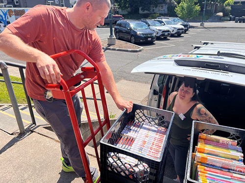 Library employee moves bins of books with a hand truck while another employee stands next to the van the bins are being loaded into