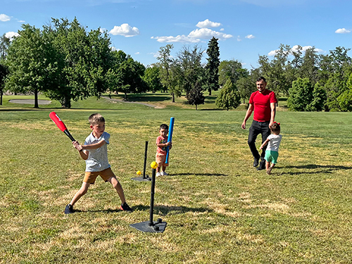 A child swings a baseball bat at a baseball on a tee while a toddler holds another bat and another toddler and a parent watch