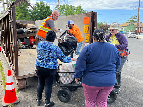 A City staff member and a volunteer lift bags of trash from a resident's wagon while another resident and volunteer watch