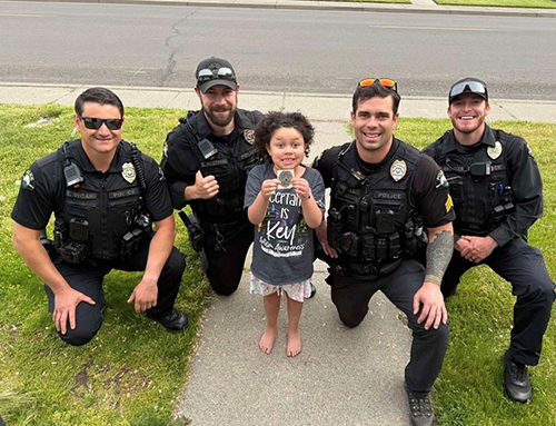 Four police officers kneel in a front yard for a photo with a child holding a sticker in the shape of a police badge