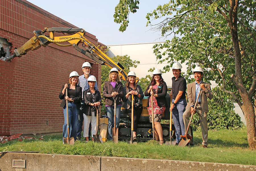Eight people wearing white hard hats hold shovels while standing on the lawn at the library in front of construction equipment